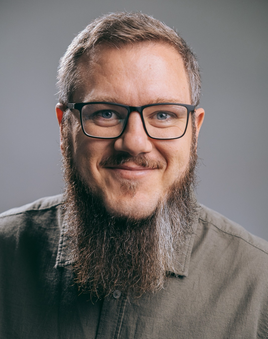 Mann mit kurzen Haaren und langem Kinnbart und Brille lächelt in die Kamera