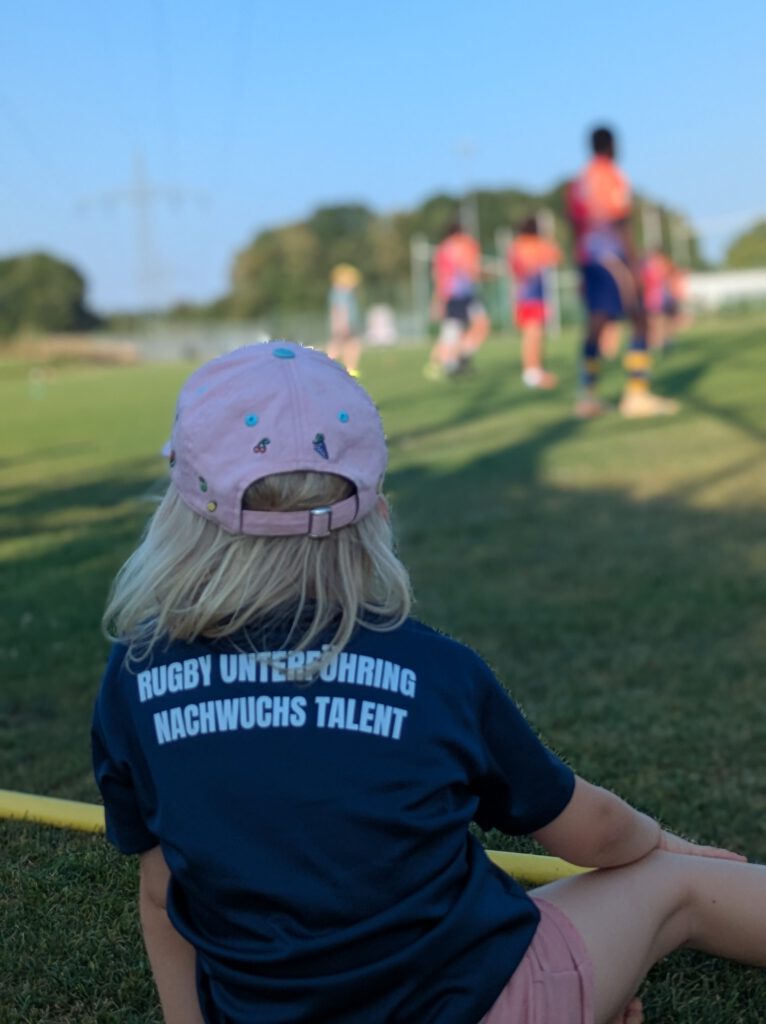 Auf dem Bild sieht man ein junges Kindergartenkind, ein blondes Mädchen mit cap am Spielfeldrand sitzen und Kinder beim Training beobachten. Sie hat auf ihrem Rücken auf dem Trikot stehen "Rugby Unterföhring Nachwuchs Talent"