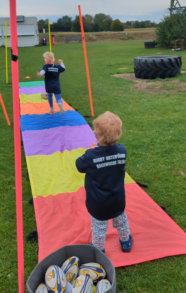 Zwei U4 Spielerinnen stehen auf einer bunten Plane in rot, gelb, lila, blau, orange und werfen einen Mini Rugby Ball in Farbfelder.